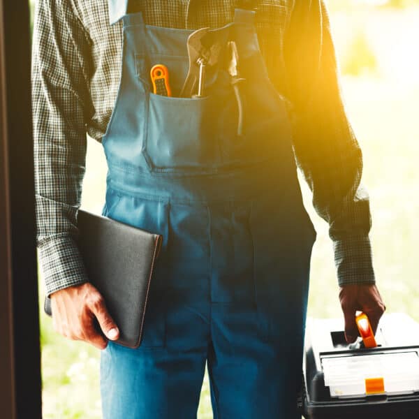 Engineer at a front door with dungarees and tools in the front pocket. He's holding a toolbox and work book