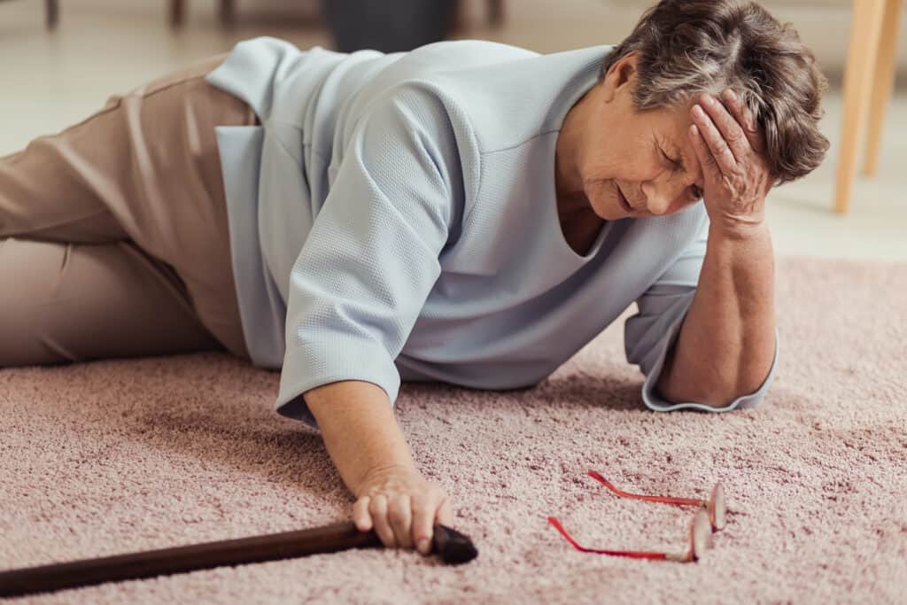 A senior woman on the floor with a headache, holding her head with one hand and her cane with the other. Her glasses are also on the carpet