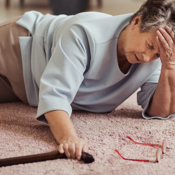A senior woman on the floor with a headache, holding her head with one hand and her cane with the other. Her glasses are also on the carpet